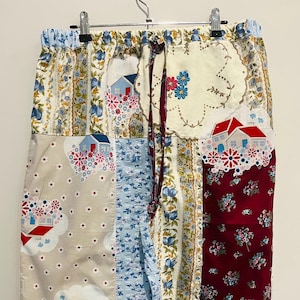 Può includere: Pantaloni patchwork con un mix di pannelli in tessuto floreale, a forma di casa e fantasia. I pantaloni presentano un punto vita con coulisse e una varietà di colori, tra cui blu, rosso, crema e beige. Il design include motivi a casa applicati e accenti floreali.