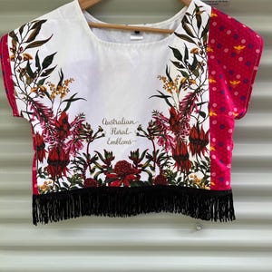 May include: White crop top with a floral design featuring red, yellow, and green accents. The right sleeve is pink with a bee pattern. The top has black fringe at the bottom and the text "Australian Floral Emblems".