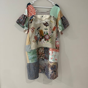 May include: A patchwork dress with a V-neck and short sleeves. The dress is made of different fabrics with floral and bird patterns. The dress has a large print of Australian birds and flowers with the word "Australia" at the bottom.