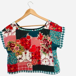 May include: A patchwork top with a variety of floral and geometric patterns in red, white, green, and black. The top is hanging on a wooden hanger. The design includes roses, polka dots, and gingham.