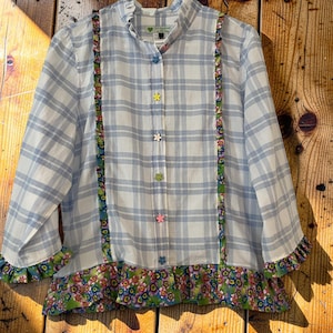 May include: A long-sleeved button-down shirt featuring a light blue and white plaid pattern. The shirt includes a ruffled collar, vertical floral-patterned trim along the front, and a floral-patterned hem. It has colourful flower-shaped buttons.