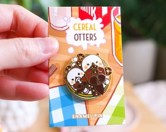 May include: Enamel pin featuring three otters in a bowl of cereal. The pin has a gold border and the words "Cereal Otters" are printed above the otters. The pin is on a card with a breakfast theme.