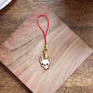 May include: A gold-colored charm with a white skull design hangs from a red string. The charm is attached to a phone case or bag.