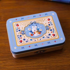 May include: A rectangular light blue metal tin featuring a central white panel with a white rabbit illustration, red strawberries, and small white flowers. The border is decorated with blue bows, red strawberry icons, and white flower motifs.