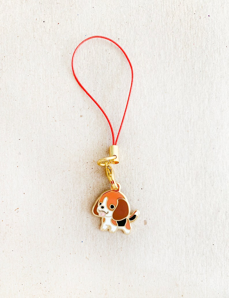 May include: A red string phone charm with a gold metal charm of a cartoon beagle dog with brown, black, and white markings.