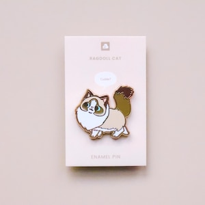 May include: Gold enamel pin featuring a cartoon Ragdoll cat with blue eyes and a speech bubble that says "Cuddle!" The pin is on a white card with the text "Ragdoll Cat" and "Enamel Pin".