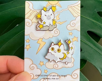 May include: Two enamel pins featuring white cats with yellow stars and a crescent moon. The pins are on a card with a blue background and clouds. The text on the card reads "Crescent Star Kittens Enamel Pin Set".