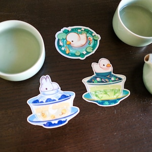 May include: Three stickers featuring cute animal illustrations. The first sticker shows a white duckling swimming in a pond with green lily pads and fish. The second sticker shows a white rabbit sitting in a blue and white teacup with a yellow moon and stars. The third sticker shows a white duckling sitting in a blue and white teacup with green water and fish.