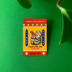 May include: A yellow and orange enamel pin featuring a cartoon tiger inside a jar with the text "Tiger Cub Jar Extra Spicy" and "I can smell just by looking at it".