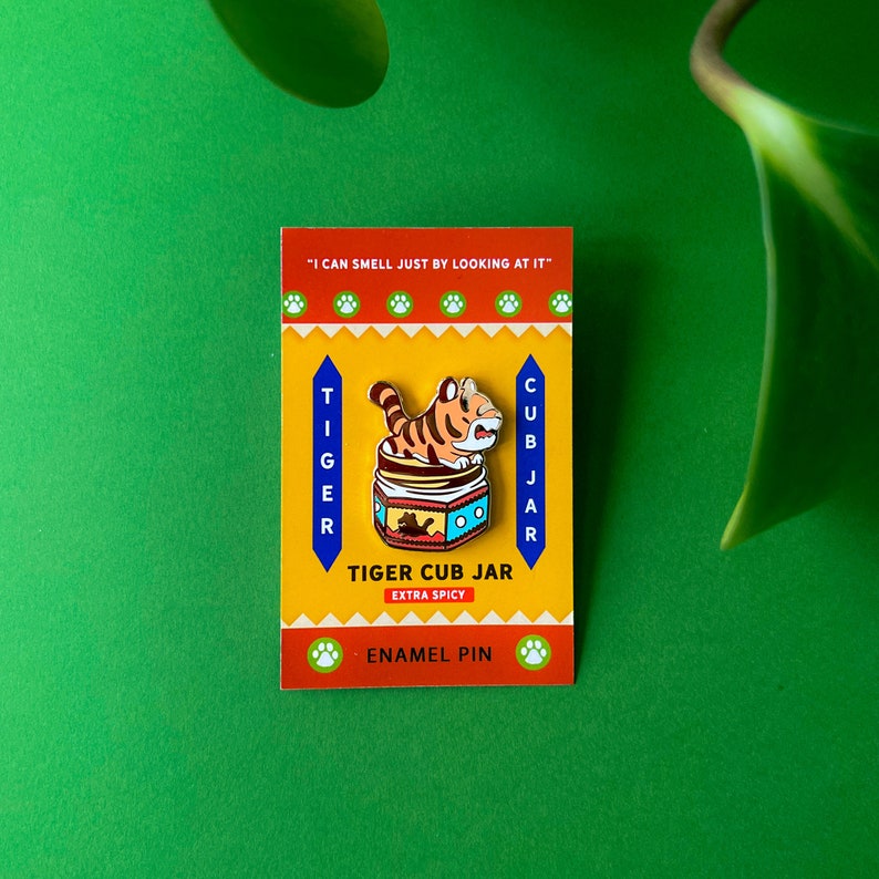 May include: A yellow and orange enamel pin featuring a cartoon tiger inside a jar with the text "Tiger Cub Jar Extra Spicy" and "I can smell just by looking at it".