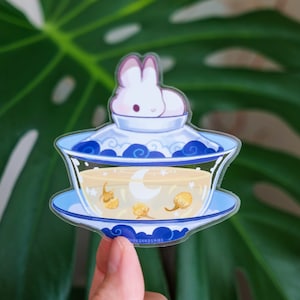 May include: A clear sticker of a white rabbit sitting in a blue and white teacup with yellow flowers and a crescent moon inside. The teacup is on a saucer with a blue and white pattern. The sticker says "thousandskies" at the bottom.