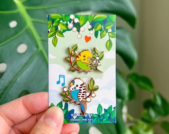 May include: A set of two enamel pins featuring colorful parakeet designs. One pin shows a yellow and green parakeet, and the other shows a blue and white parakeet. The pins are on a card with the text "BFF Buggies ENAMEL PIN SET".