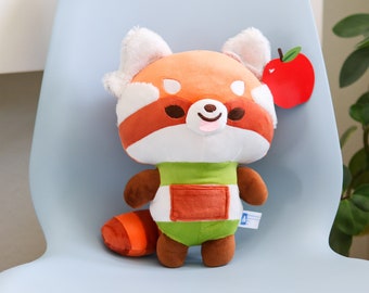 May include: A plush red panda toy wearing a green and orange outfit with a pocket. The toy has a white face with black eyes and a pink tongue sticking out. A red apple is in the background.