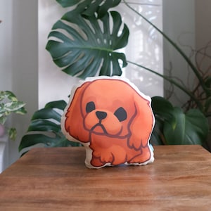 May include: A plush pillow shaped like a cartoon Cavalier King Charles Spaniel dog. The dog is brown and has a white patch on its chest. The pillow is made of soft fabric and has a cute, cartoonish design.