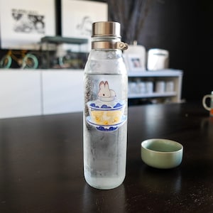 May include: A clear glass bottle with a silver lid and a sticker of a white rabbit sitting in a blue and white bowl. The bottle is sitting on a dark wooden table.