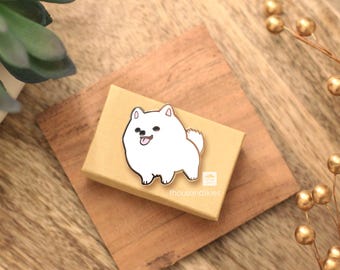 May include: White enamel pin of a cartoon dog with a pink tongue sticking out. The pin is on a brown box with the text "theoutboundclub" on it.