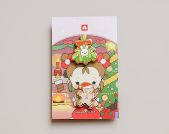 May include: A set of two enamel pins featuring a girl with a gingerbread man and a mistletoe with a bunny. The pins are on a card with a red and green Christmas scene and the text "Enamel Pin Set".