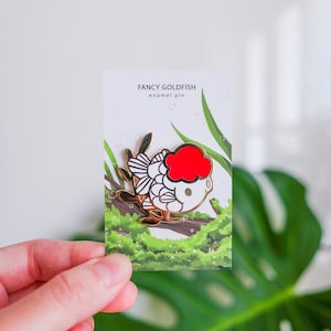 May include: Gold enamel pin of a white goldfish with a red fin, on a card that says "Fancy Goldfish enamel pin".