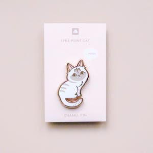 May include: A white and gray enamel pin of a lynx point cat with a speech bubble that says "meows". The pin is on a white card with the text "LYNX POINT CAT" and "ENAMEL PIN".