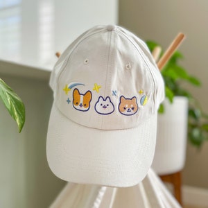 May include: White baseball cap with embroidered cartoon dogs and stars. The dogs are a corgi, a pug, and a shiba inu.