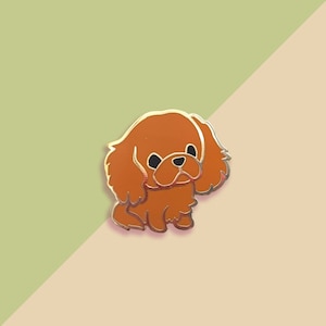 May include: A gold-colored enamel pin featuring a cartoon illustration of a brown Cavalier King Charles Spaniel dog with black eyes.