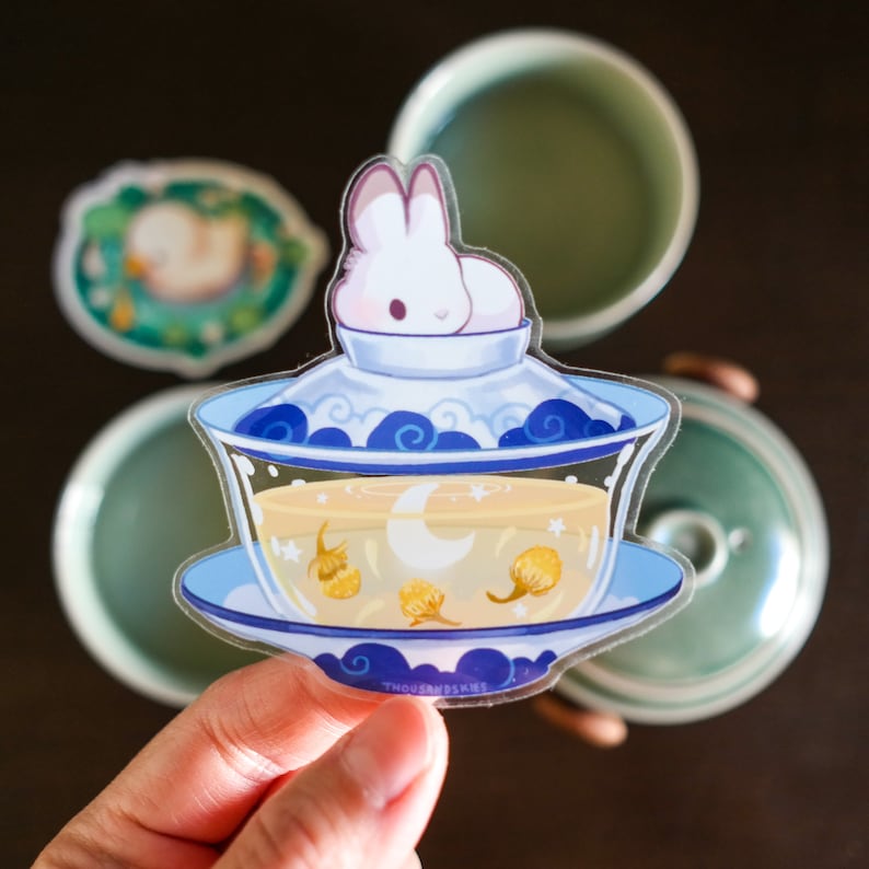 May include: A white rabbit sticker with blue eyes is sitting on top of a blue and white teacup with a clear liquid inside. The teacup is on a white sticker with a blue border and the text "THOUSANDSKIES" in blue.
