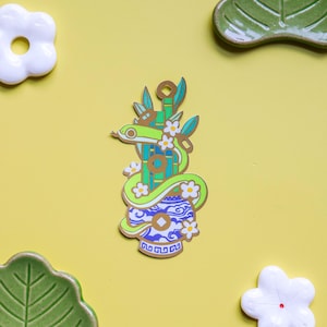 Peut inclure: Épingle décorative représentant un serpent vert enroulé autour de tiges de bambou et d'un vase bleu et blanc. L'épingle est ornée de petites fleurs blanches et de détails dorés, sur fond jaune avec des éléments décoratifs blancs et verts.