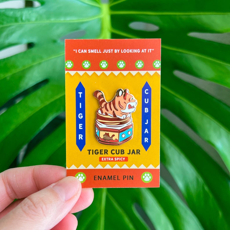 May include: A yellow enamel pin with a cartoon tiger cub inside a jar. The jar is labeled "Tiger Cub Jar" and "Extra Spicy". The text "I can smell just by looking at it" is at the top of the pin.
