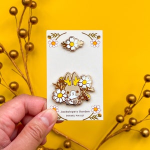 May include: A set of two enamel pins featuring a brown bunny with yellow ears surrounded by white daisies and a single daisy. The pins are on a white card with the text "Jackalope's Garden Enamel Pin Set".