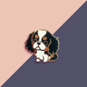 May include: Gold enamel pin featuring a cartoon Cavalier King Charles Spaniel with black, white and brown fur. The dog is looking to the left.