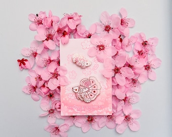 Può includere: Un set di due spille smaltate Sakura Halfmoon Betta su una carta rosa, circondate da fiori di ciliegio rosa. Le spille presentano un design dettagliato di un pesce betta. La carta recita "Sakura Halfmoon Betta Enamel Pin Set".