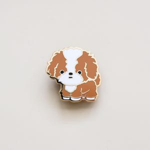 May include: A gold-tone enamel pin featuring a cartoon illustration of a brown and white Shih Tzu dog. The dog is standing on all four paws with its tongue sticking out.