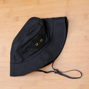 May include: Black bucket hat with a cat face applique. The cat face is made of black fabric with gold-colored eyes. The hat has a black adjustable chin strap. The hat is made of a soft fabric.