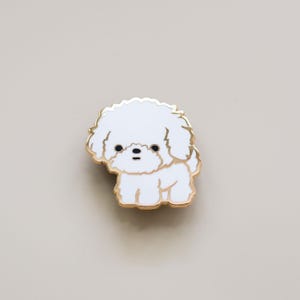 May include: A white enamel pin of a small, fluffy white dog with black eyes. The dog is standing on all four paws and has a gold outline.