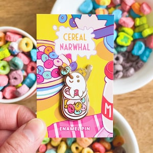 May include: A gold enamel pin featuring a cartoon narwhal in a bowl of cereal. The pin says "Cereal Narwhal" and "Enamel Pin".