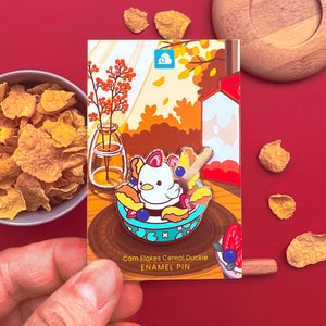 Peut inclure: Épinglette en émail représentant un petit canard de dessin animé dans un bol de céréales avec des flocons de maïs, des fraises et des myrtilles. L'épingle est sur une carte avec le texte "Corn Flakes Cereal Duckie Enamel Pin".