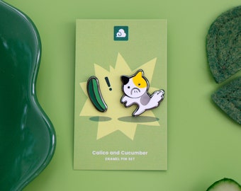 May include: A set of two enamel pins featuring a calico cat and a cucumber. The cat is white with black and yellow markings and is running away from the cucumber. The pins are on a white card with the text "Calico and Cucumber Enamel Pin Set".