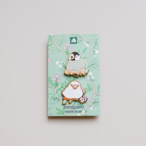 May include: A set of two enamel pins featuring a Java Sparrow bird. The pins are gold-toned with white and gray enamel. The pins are on a card with a floral design and the text "Java Sparrow Enamel Pin Set".