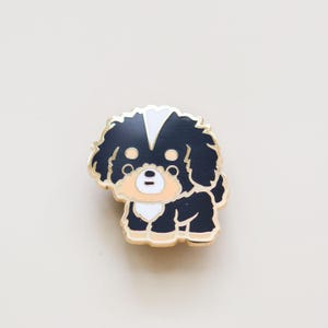 May include: Gold-tone enamel pin featuring a black and white cartoon dog with a white patch on its head. The dog has a black nose and brown eyes.