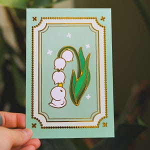 May include: A mint green card with a gold border and a lily of the valley illustration. The illustration features white bell-shaped flowers, green leaves, and a gold outline. Small white flower accents are scattered around the image.