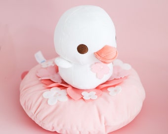 May include: A white plush duck with brown eyes and a pink beak sitting on a pink plush cushion with white flower accents.