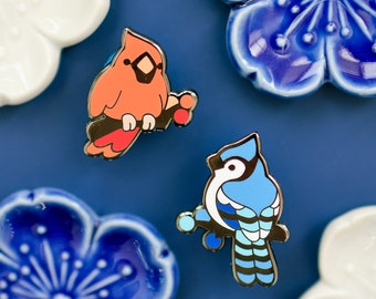 Op de afbeelding: Twee emaillepins met een rode kardinaal en een blauwe gaaien. De kardinaal zit op een tak met rode bessen. De blauwe gaaien zit op een tak met een blauw-wit gestreept patroon. Beide pins hebben een zilveren afwerking.