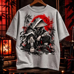 May include: A light grey short-sleeved t-shirt featuring a graphic print of a crouching figure wearing a conical hat and dark robes, holding a katana. The design includes a large red sun, black bamboo silhouettes, and intricate geometric patterns.