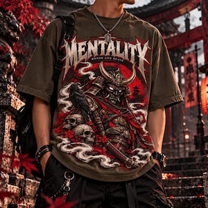 May include: An olive green t-shirt featuring a graphic print of a samurai in armour with a horned helmet, skulls, and a crow. The text MENTALITY and HONOR AND DEATH is printed in a stylised font above the samurai. The design includes red accents.