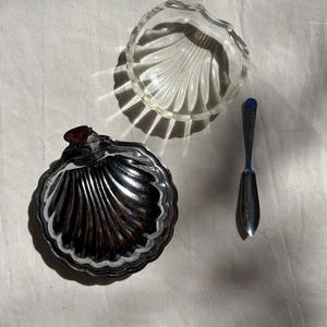 Op de afbeelding: Een heldere glazen schaal in de vorm van een schelp, een bijpassend deksel van zilverkleurig metaal in de vorm van een schelp en een klein zilverkleurig metalen smeermesje. De glazen schaal heeft een geschulpte rand en een geribbelde textuur.