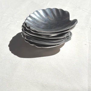 May include: A stack of six silver-toned metal dishes shaped like scallop shells. Each dish features a fluted, fan-like design with a smooth, reflective surface. The dishes are nested together, creating a layered appearance with visible ridges.