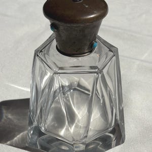 Op de afbeelding: Een parfumflesje van helder glas met een gefacetteerd, geometrisch lichaam en een donkere metalen dop. De dop is versierd met kleine, ronde, fel turquoise gekleurde cabochons die rond de bovenkant en zijkanten van het deksel zijn geplaatst.