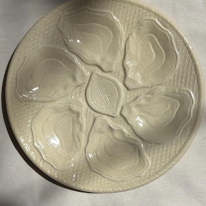 May include: A round, cream-colored ceramic oyster plate featuring five molded indentations shaped like oyster shells arranged in a circle around a central leaf-like motif. The surface has a textured, cross-hatched pattern throughout.