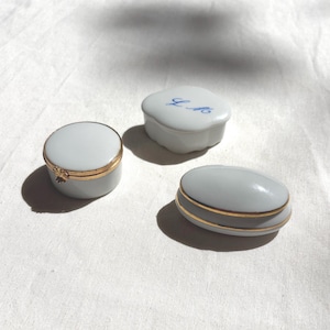 May include: Three small porcelain trinket boxes with smooth white surfaces and gold-toned metal rims. One box is round, one is oval, and one is square with rounded corners featuring the blue cursive initials L.M. on the lid.