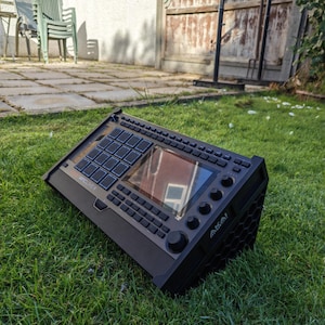 May include: A black Akai MPC Live II music production workstation featuring a central touchscreen, a grid of sixteen backlit drum pads, multiple control knobs, and function buttons. The device has a textured chassis with a honeycomb side pattern.